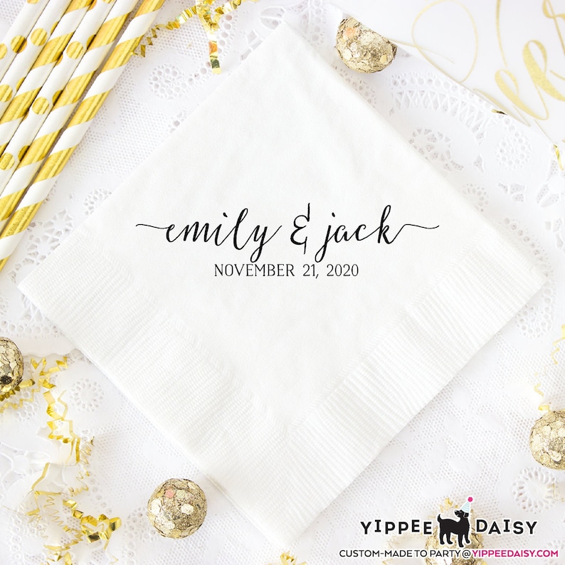 May include: White paper napkin with black calligraphy text that reads "emily & jack" and "NOVEMBER 21, 2020". The napkin is on a white lace tablecloth with gold confetti and gold-colored ornaments.