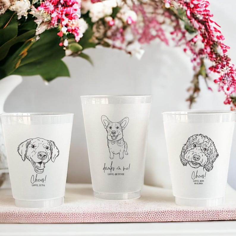May include: Three frosted plastic cups with black line drawings of dogs. One cup has a Labrador, another a Corgi, and the third a Goldendoodle. Text includes "Cheers!" and "drinks on me!" with names below. Pink flowers are in the background.