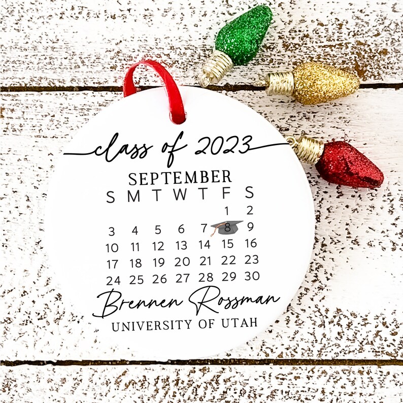May include: White round bauble with black text that reads "class of 2023", "SEPTEMBER", "SMTWTFS", "1", "2", "3", "4", "5", "6", "7", "8", "9", "10", "11", "12", "13", "14", "15", "16", "17", "18", "19", "20", "21", "22", "23", "24", "25", "26", "27", "28", "29", "30", "Brennen Rossman", "UNIVERSITY OF UTAH". A small black graduation cap icon is next to the number "8".