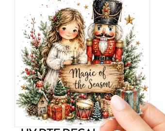 Op de afbeelding: Een UV DTF-sticker met een meisje in een witte jurk dat een kaars vasthoudt naast een notenkraker, met een houten bord met de tekst Magic of the Season, omringd door kerstbomen, cadeaus, een trommel en sterren op een doorzichtige achtergrond.