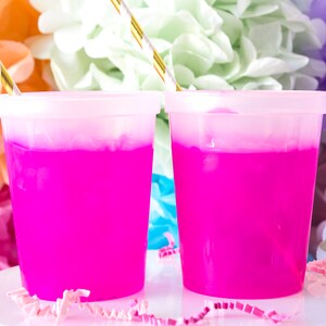 May include: Two pink plastic cups with clear liquid and gold straws. The cups are on a white surface with pink and white confetti.