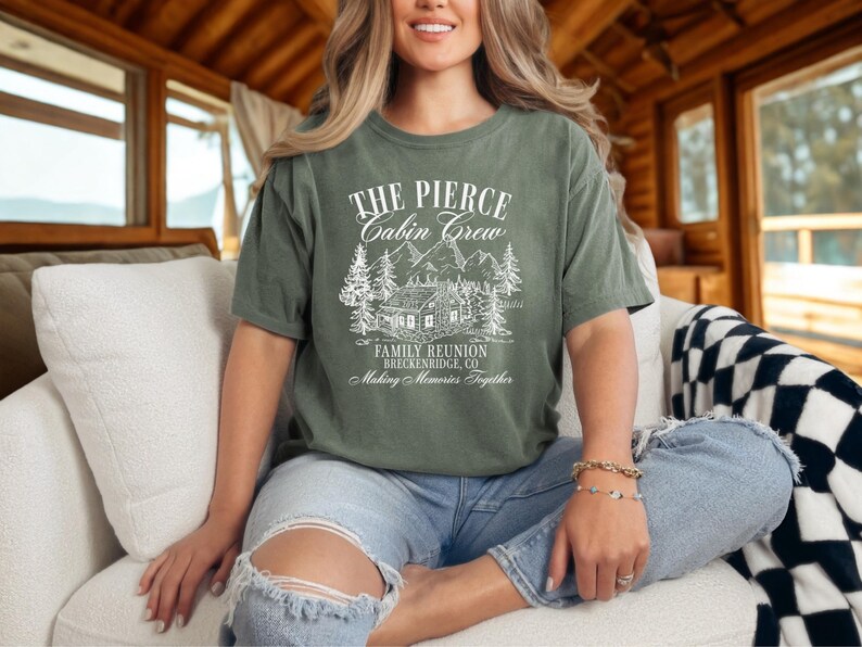 Pu&ograve; includere: T-shirt verde oliva con testo e illustrazione bianchi. Il testo recita "THE PIERCE CABIN CREW FAMILY REUNION BRECKENRIDGE, CO Making Memories Together." L'illustrazione raffigura una baita, alberi e montagne. La maglietta &egrave; indossata.
