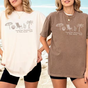 May include: Two oversized t-shirts, one cream and one brown, featuring a beach-themed graphic. The graphic includes an umbrella, beach ball, chair, seashell, and palm tree, with the text "Mackenzie's Bachelorette St. Pete, Florida - 2025".