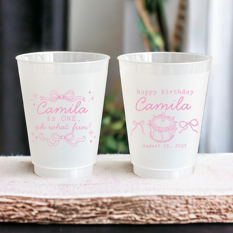 May include: Two white plastic cups with pink text and designs. One cup says "Camila is ONE, oh what fun!" The other cup says "happy birthday Camila August 23, 2025".