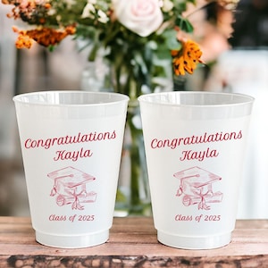 Puede incluir: Dos vasos de pl&aacute;stico blanco con texto rojo y un dise&ntilde;o de gorro de graduaci&oacute;n. El texto en los vasos dice "Felicidades Kayla Clase de 2025".