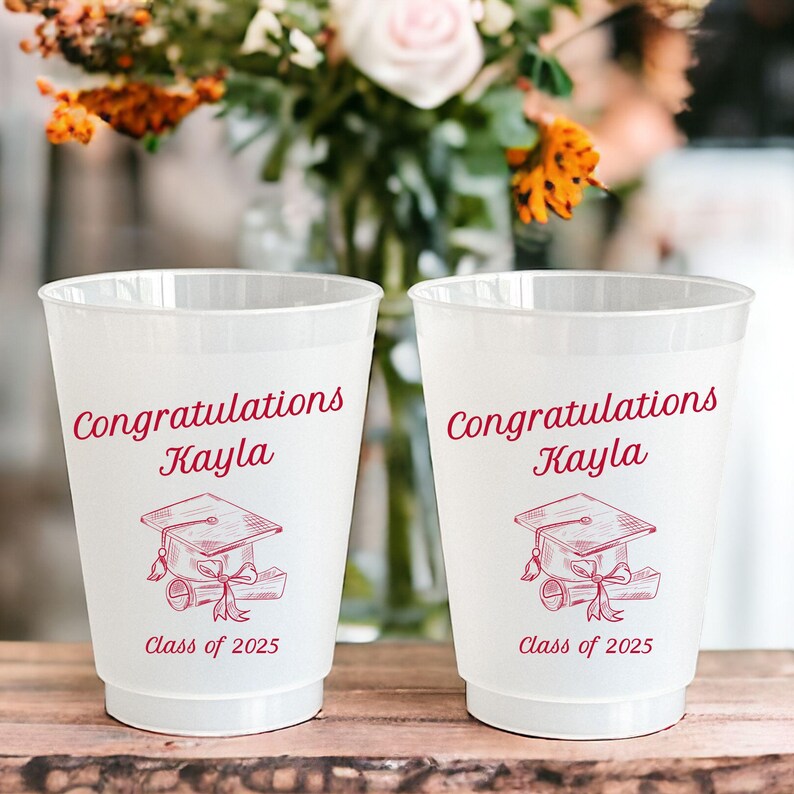 Puede incluir: Dos vasos de pl&aacute;stico blanco con texto rojo y un dise&ntilde;o de gorro de graduaci&oacute;n. El texto en los vasos dice "Felicidades Kayla Clase de 2025".