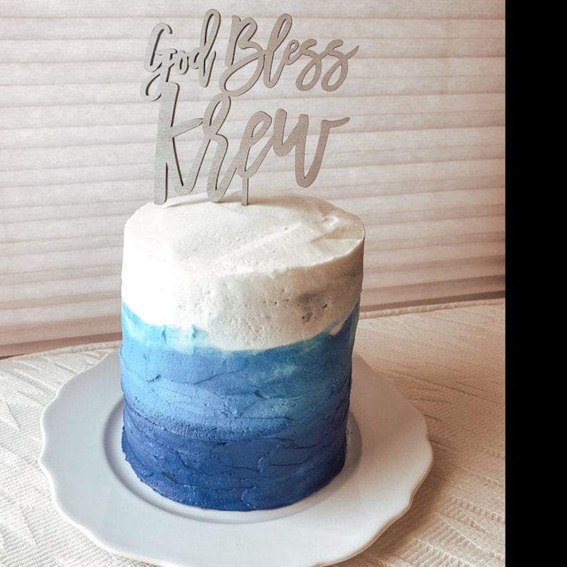 May include: A white and blue ombre cake with a wooden cake topper that says "God Bless Crew".