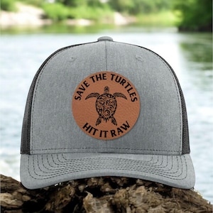 May include: A gray and black trucker hat with a brown leather patch that says "Save the Turtles Hit it Raw" and features a black silhouette of a turtle with a floral design.
