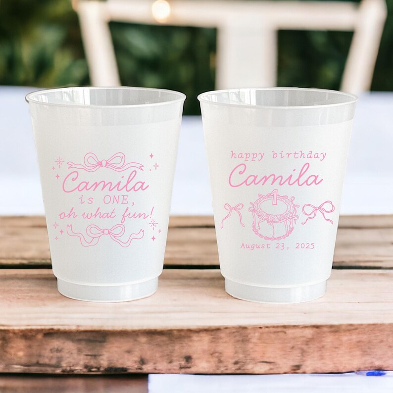 May include: Two pink disposable cups with a white background. One cup says "Camila is ONE, oh what fun!" with a pink bow and stars. The other cup says "happy birthday Camila" with a pink cake and bows. The date August 23, 2025 is printed below the text.