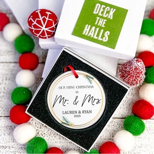 Op de afbeelding: Witte kerstbal van keramiek met gouden rand en groene dennentakken. De kerstbal heeft de tekst "Our First Christmas as Mr. & Mrs. Lauren & Ryan 2023".