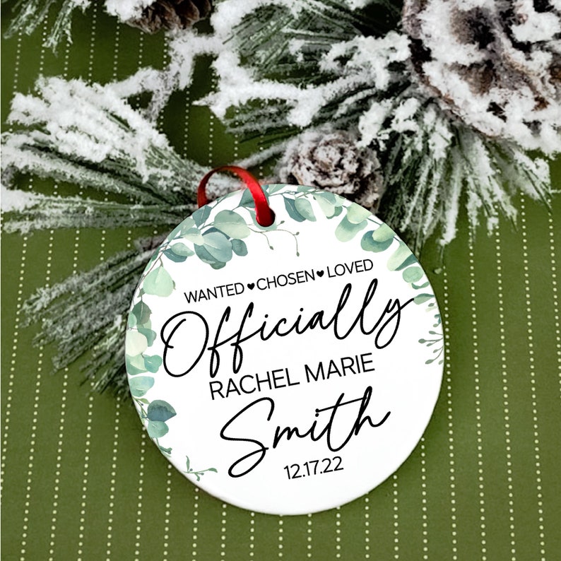 Op de afbeelding: Ronde witte ornament met een groen eucalyptusbladeren ontwerp. De ornament heeft zwarte tekst die "Wanted &hearts; Chosen &hearts; Loved Officially Rachel Marie Smith 12.17.22" luidt.