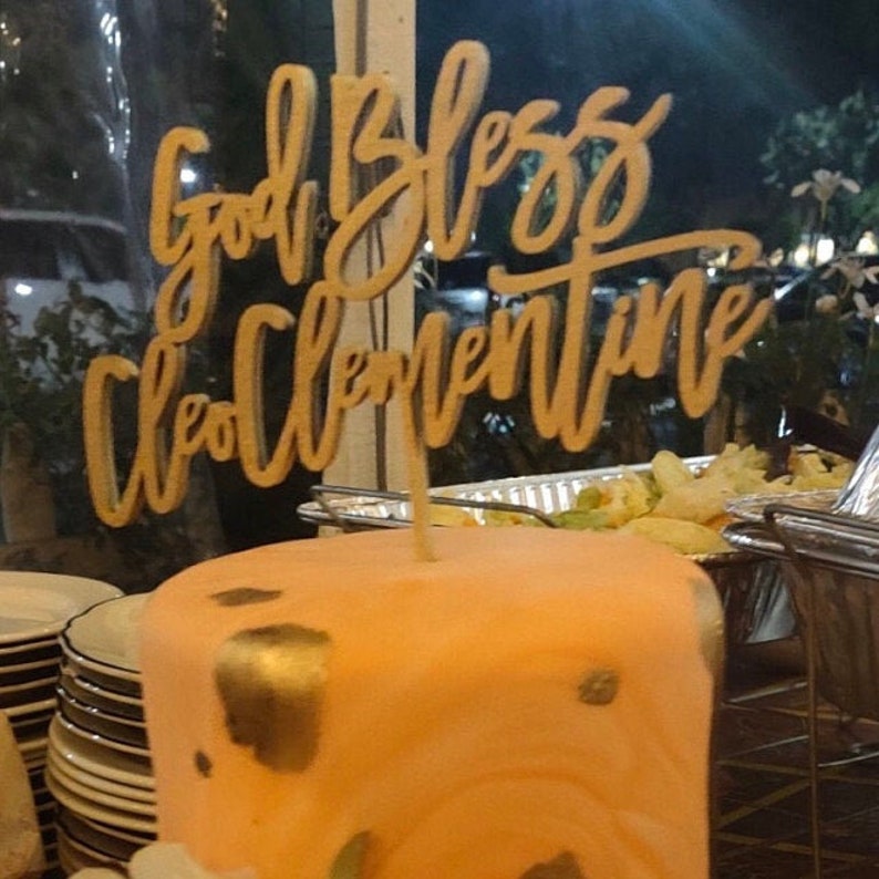 May include: A peach-colored cake with gold accents and a wooden cake topper that says "God Bless Clementine".