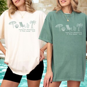 May include: Two oversized t-shirts, one cream and one sage green, featuring a beach-themed graphic and text. The graphic includes an umbrella, seashell, chair, and palm tree. The text reads "Mackenzie's Bachelorette St. Pete, Florida - 2025."