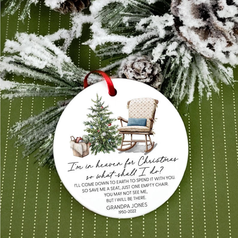 May include: A white ceramic Christmas bauble featuring a rocking chair, a Christmas tree, and a basket of gifts. The bauble is inscribed with the text "I'm in heaven for Christmas so what shall I do? I'll come down to earth to spend it with you. So save me a seat, just one empty chair. You may not see me, but I will be there. Grandpa Jones 1950-2022".