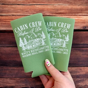 May include: Two green can coolers with white text that reads "CABIN CREW Before I Do" above a cabin and mountain graphic. The text "MACY'S BACHELORETTE HOCKING HILLS, OHIO | 2025" is printed below the graphic.