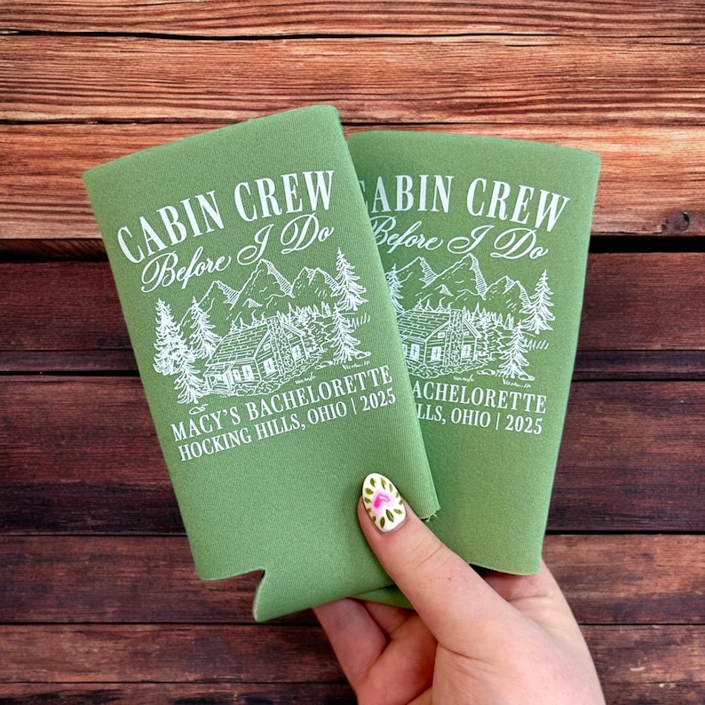 May include: Two green can coolers with white text that reads "CABIN CREW Before I Do" above a cabin and mountain graphic. The text "MACY'S BACHELORETTE HOCKING HILLS, OHIO | 2025" is printed below the graphic.