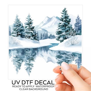 May include: A clear UV DTF decal featuring a watercolor winter landscape with snow-covered pine trees, mountains, and a reflective river. The design includes white snow accents and blue-toned foliage. Text at the bottom reads UV DTF DECAL.