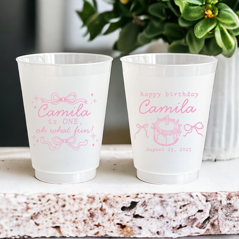 May include: Two white plastic cups with pink text and designs. One cup says "Camila is ONE, oh what fun!" and the other says "happy birthday Camila" with a cake and the date "August 23, 2025".