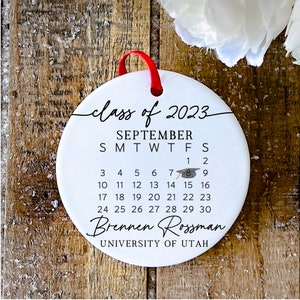 May include: White ceramic ornament with a black calendar for September 2023, featuring a graduation cap icon on the 8th. The text "class of 2023" is written in a cursive font at the top. Below the calendar, the text "Brennen Rossman" and "UNIVERSITY OF UTAH" are written in black.