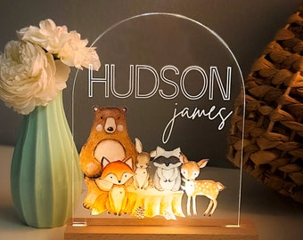 May include: A clear acrylic night light with a wooden base. The light features a woodland scene with a bear, fox, rabbit, raccoon, and deer. The names "HUDSON james" are written on the light.