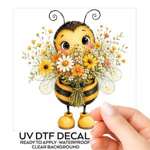 May include: A UV DTF decal featuring a cartoon bee with black and yellow stripes, large eyes, and yellow boots, holding a large bouquet of yellow, orange, and white wildflowers. The text below reads UV DTF DECAL READY TO APPLY WATERPROOF CLEAR BACKGROUND.