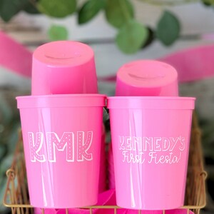 May include: Two pink plastic cups with white lettering. The cup on the left says "KMK" and the cup on the right says "Kennedy's First Fiesta!"