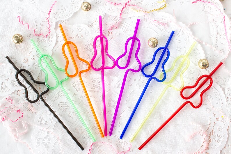 May include: A set of eight colourful plastic straws with a unique bend in the shape of a heart. The straws are arranged in a fan shape on a white lace background.