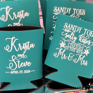 May include: Teal blue can coolers with white text. One cooler says "Sandy Toes and Salty Kisses Cheers to the New Mr. & Mrs." The other cooler says "Krista and Steve - April 27, 2019 - "