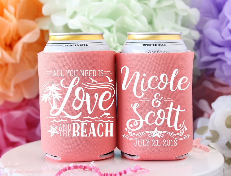 May include: Two pink foam can coolers with white text. One cooler says "All you need is Love and the Beach" with a palm tree, ocean waves, and a starfish. The other cooler says "Nicole & Scott" with a starfish and the date "July 21, 2018".