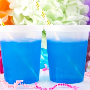 May include: Two clear plastic cups filled with blue liquid and topped with white lids.  The cups have gold and white striped straws inserted in them. The cups are sitting on a white surface with pink and white confetti.