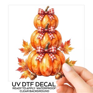 May include: A UV DTF decal featuring a vertical stack of three orange pumpkins, each adorned with a red and white gingham bow. The design is accented with colorful autumn maple leaves and small brown acorns at the base, set against a clear background.