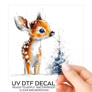 May include: A UV DTF decal featuring a watercolor-style illustration of a young deer with brown fur and white spots standing next to a small blue pine tree. The text below reads UV DTF DECAL READY TO APPLY WATERPROOF CLEAR BACKGROUND.