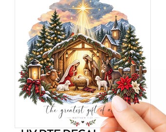 Op de afbeelding: Een UV DTF-sticker met een kerststalscène met een stal, figuren, dieren en een heldere ster erboven. De tekst luidt "the greatest gift of all" en "MATTHEW 1:21". Het ontwerp bevat besneeuwde dennenbomen, een lantaarn en kerststerren.