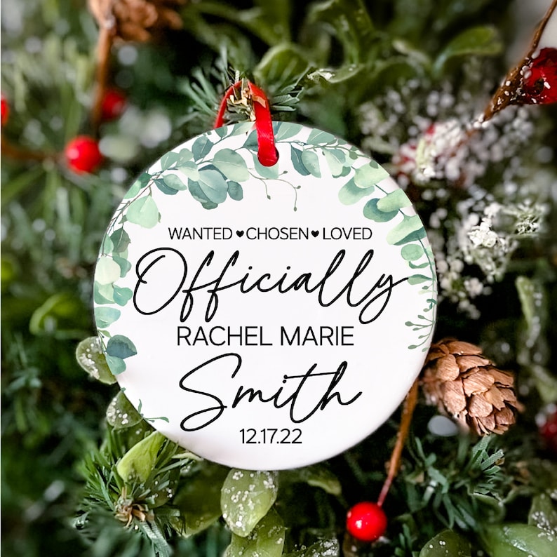 Op de afbeelding: Ronde witte kerstbal met een ontwerp van groene eucalyptusbladeren. De bal heeft de tekst "Wanted &hearts; Chosen &hearts; Loved Officially Rachel Marie Smith 12.17.22" erop gedrukt.