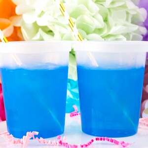 Może przedstawiać: Dwa przezroczyste plastikowe kubki wypełnione niebieskim płynem i przykryte białymi pokrywkami. Słomki w paski w kolorze złotym i białym są włożone do kubk&oacute;w.