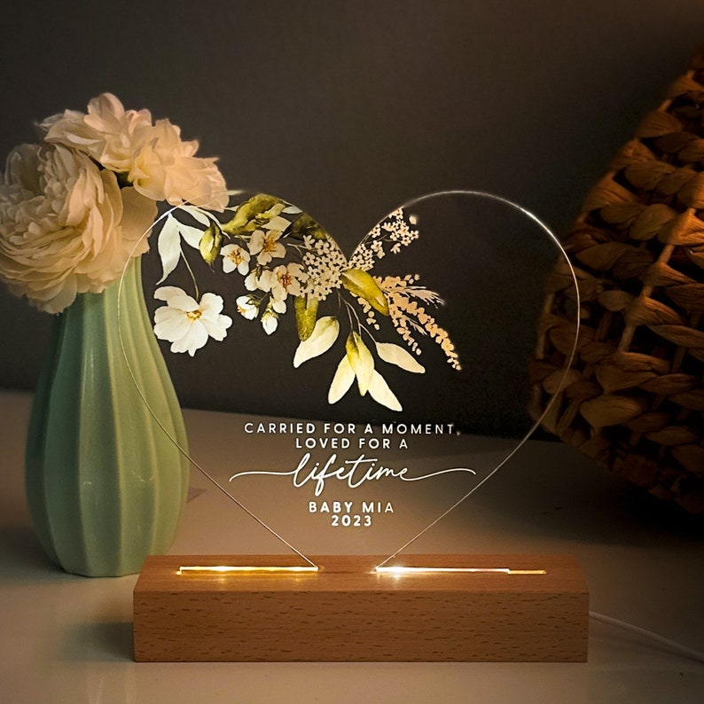May include: A heart-shaped acrylic sign with a floral design and the text "Carried for a moment, loved for a lifetime. Baby Mia 2023". The sign is illuminated with warm white LED lights and sits on a wooden base.