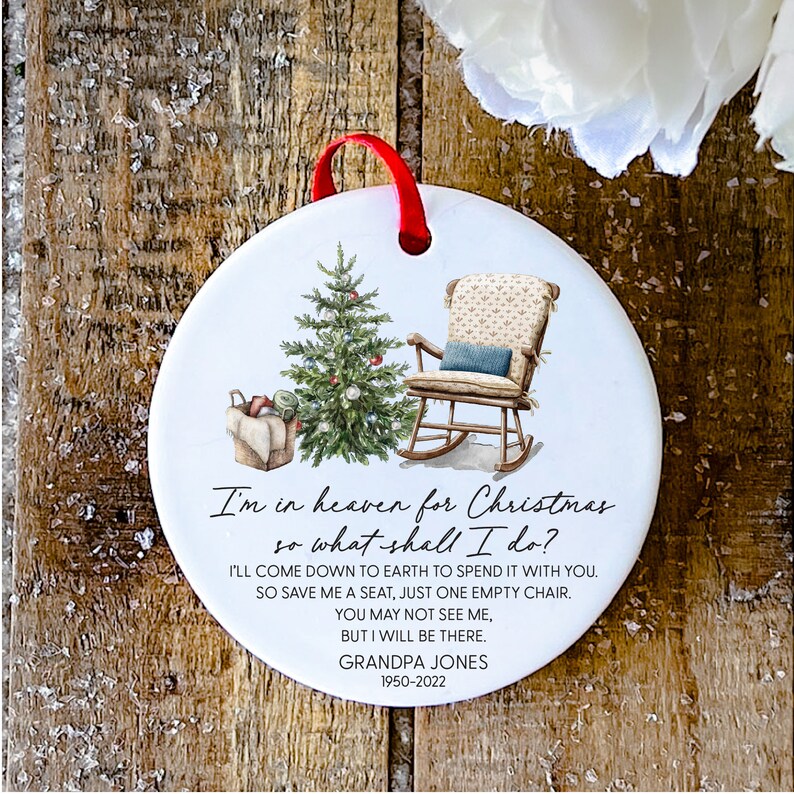 May include: White ceramic Christmas bauble featuring a hand-painted image of a rocking chair, a Christmas tree, and a basket of gifts. The ornament includes the text "I'm in heaven for Christmas so what shall I do? I'll come down to earth to spend it with you. So save me a seat, just one empty chair. You may not see me, but I will be there. Grandpa Jones 1950-2022".