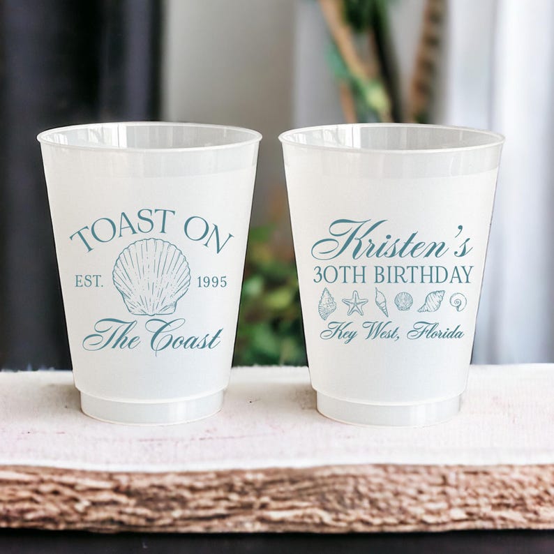 K&ouml;nnte beinhalten: Zwei wei&szlig;e Plastikbecher mit blauem Text und Grafiken. Der linke Becher sagt "Toast On Est. 1995 The Coast" mit einer Muschelgrafik. Der rechte Becher sagt "Kristen's 30th Birthday Key West, Florida" mit Muschel- und Seestern-Grafiken.