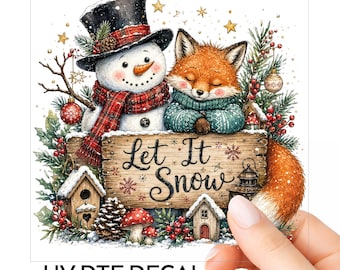 Op de afbeelding: Een UV DTF-sticker met een sneeuwpop in een zwarte hoge hoed en rood geruite sjaal naast een vos in een blauwgroene trui. Ze staan achter een houten bord met de tekst Let It Snow, omringd door dennentakken, rode bessen en kleine vogelhuisjes.