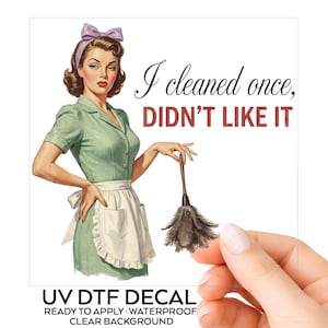 May include: A UV DTF decal featuring a vintage-style illustration of a person in a green dress, white apron, and purple hair bow holding a feather duster. The text reads I cleaned once, DIDN'T LIKE IT. The decal has a clear background.