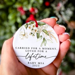 May include: White ceramic ornament with a floral design and the text "Carried for a moment, loved for a lifetime. Baby Mia 2023".