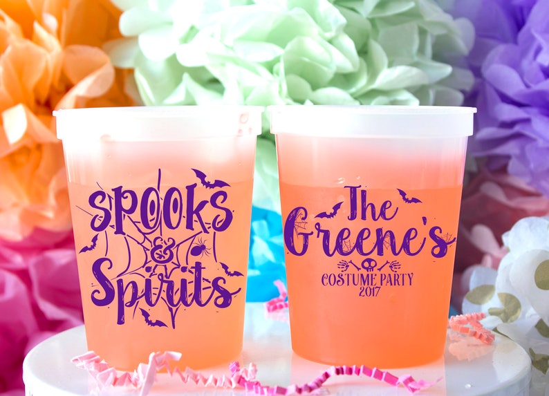 Może przedstawiać: Dwa pomarańczowe plastikowe kubki z fioletowym tekstem. Jeden kubek ma napis "Spooks & Spirits" z pajęczyną i nietoperzami. Drugi kubek ma napis "The Greene's Costume Party 2017".