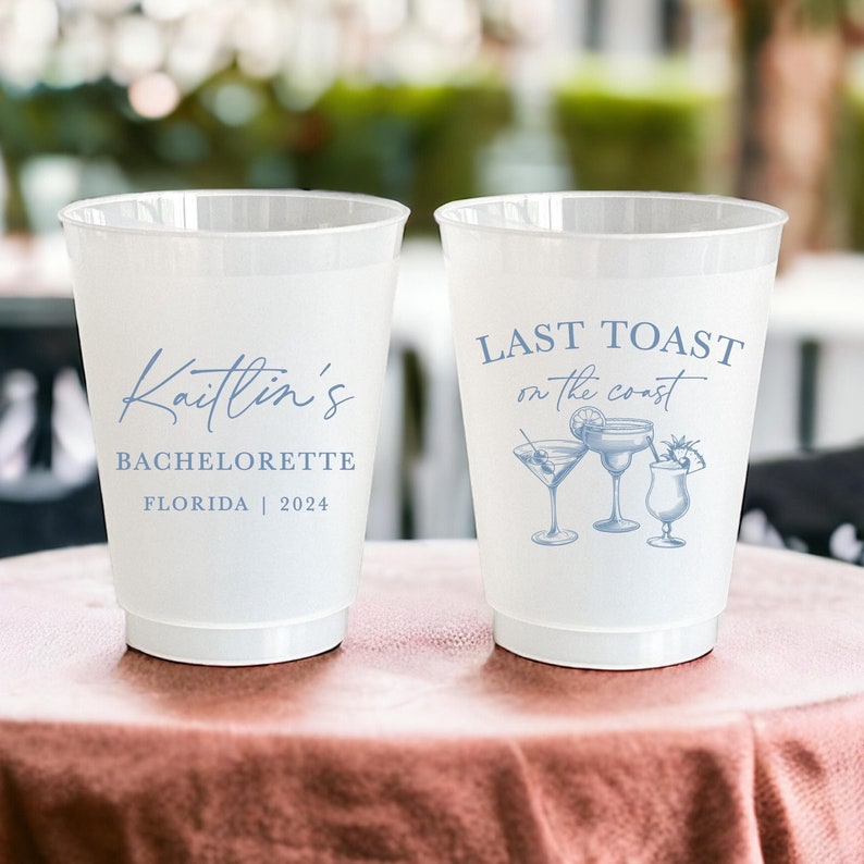 May include: Two white plastic cups with blue text and designs. One cup says "Kaitlin's Bachelorette Florida | 2024". The other cup says "Last Toast on the Coast" with a drawing of three cocktails.