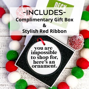 K&ouml;nnte beinhalten: Wei&szlig;er Keramik-Weihnachtsbaumschmuck mit schwarzem Text, der "Du bist unm&ouml;glich zu beschenken, hier ist ein Ornament" lautet. Der Schmuck befindet sich in einer wei&szlig;en Geschenkbox mit rotem Band. Die Box ist von wei&szlig;en, roten und gr&uuml;nen Bommeln umgeben.