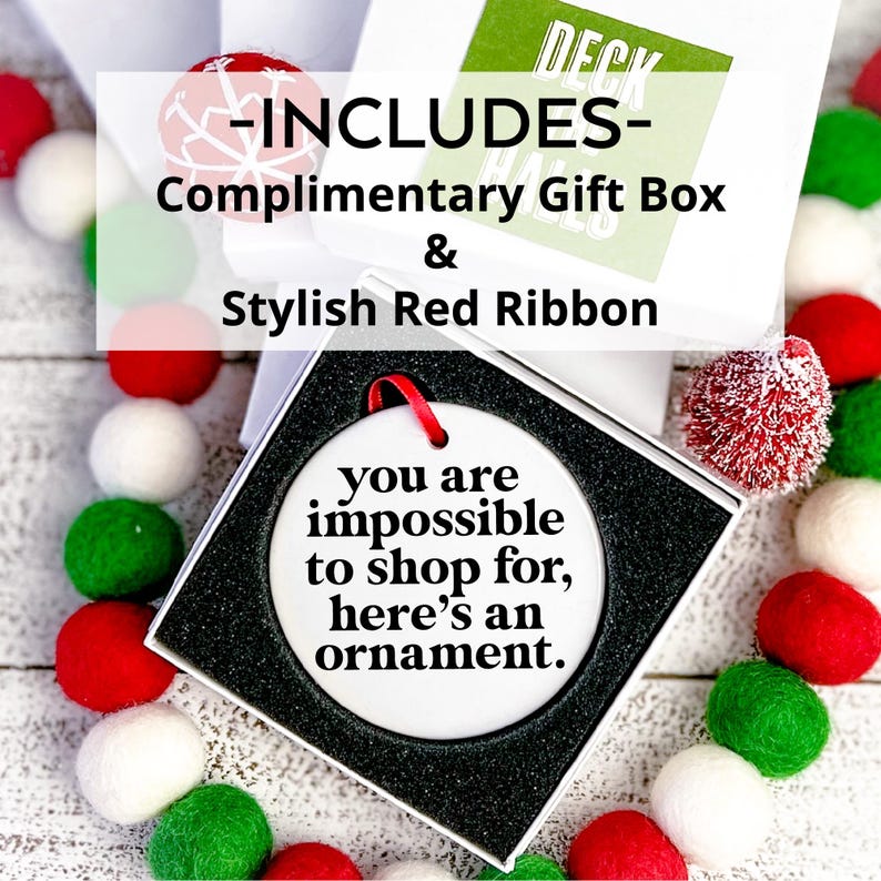 K&ouml;nnte beinhalten: Wei&szlig;er Keramik-Weihnachtsbaumschmuck mit schwarzem Text, der "Du bist unm&ouml;glich zu beschenken, hier ist ein Ornament" lautet. Der Schmuck befindet sich in einer wei&szlig;en Geschenkbox mit rotem Band. Die Box ist von wei&szlig;en, roten und gr&uuml;nen Bommeln umgeben.