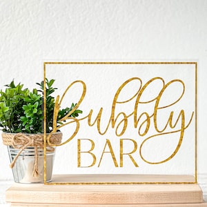 May include: A clear acrylic sign with a gold glitter finish. The sign reads "Bubbly Bar" in a gold cursive font.