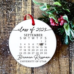 May include: White round bauble with a black calendar for September 2023, the text "class of 2023", a graduation cap icon, and the name "Brennen Rossman" with the text "UNIVERSITY OF UTAH" underneath.