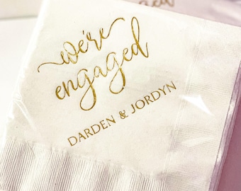 Könnte beinhalten: Weiße Papierservietten mit goldfarbener Folienbeschriftung mit dem Text "we're engaged" und "DARDEN & JORDYN".