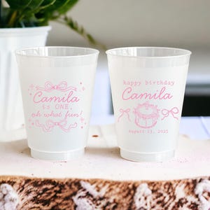 May include: Two white plastic cups with pink text and illustrations. One cup says "Camila is ONE, oh what fun!" and the other says "happy birthday Camila" with a cake and the date "August 23, 2025".