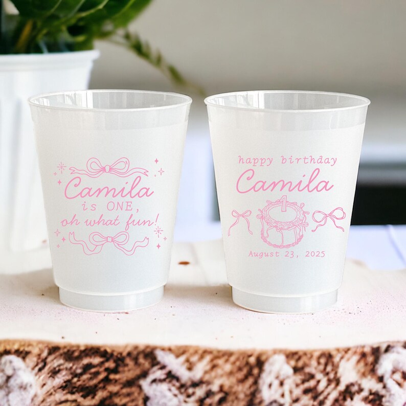May include: Two white plastic cups with pink text and illustrations. One cup says "Camila is ONE, oh what fun!" and the other says "happy birthday Camila" with a cake and the date "August 23, 2025".